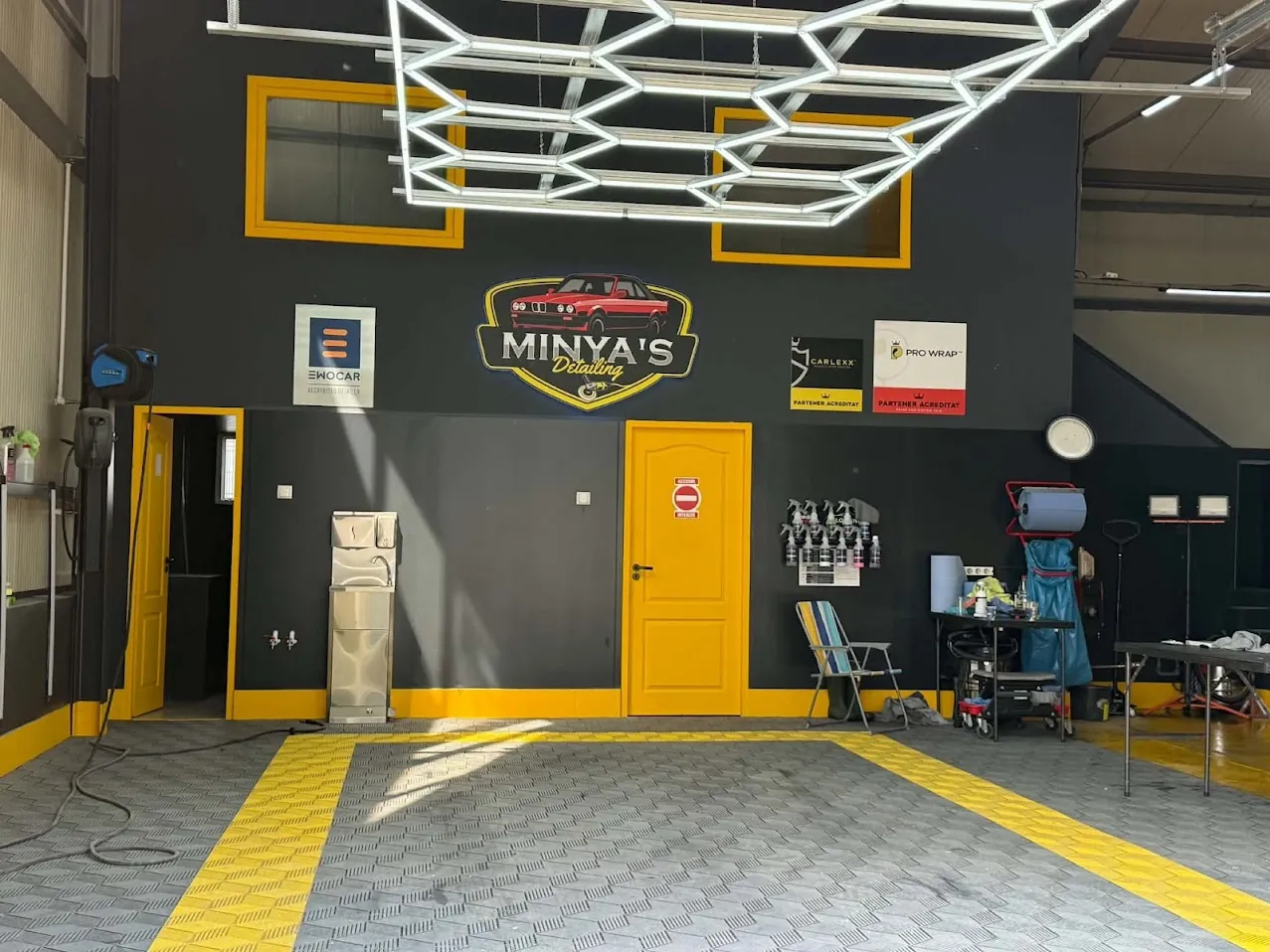 Atelierul MINYA'S Detailing din Livezeni: grila de lumini hexagonale, sigla, pardoseala gri cu benzi galbene